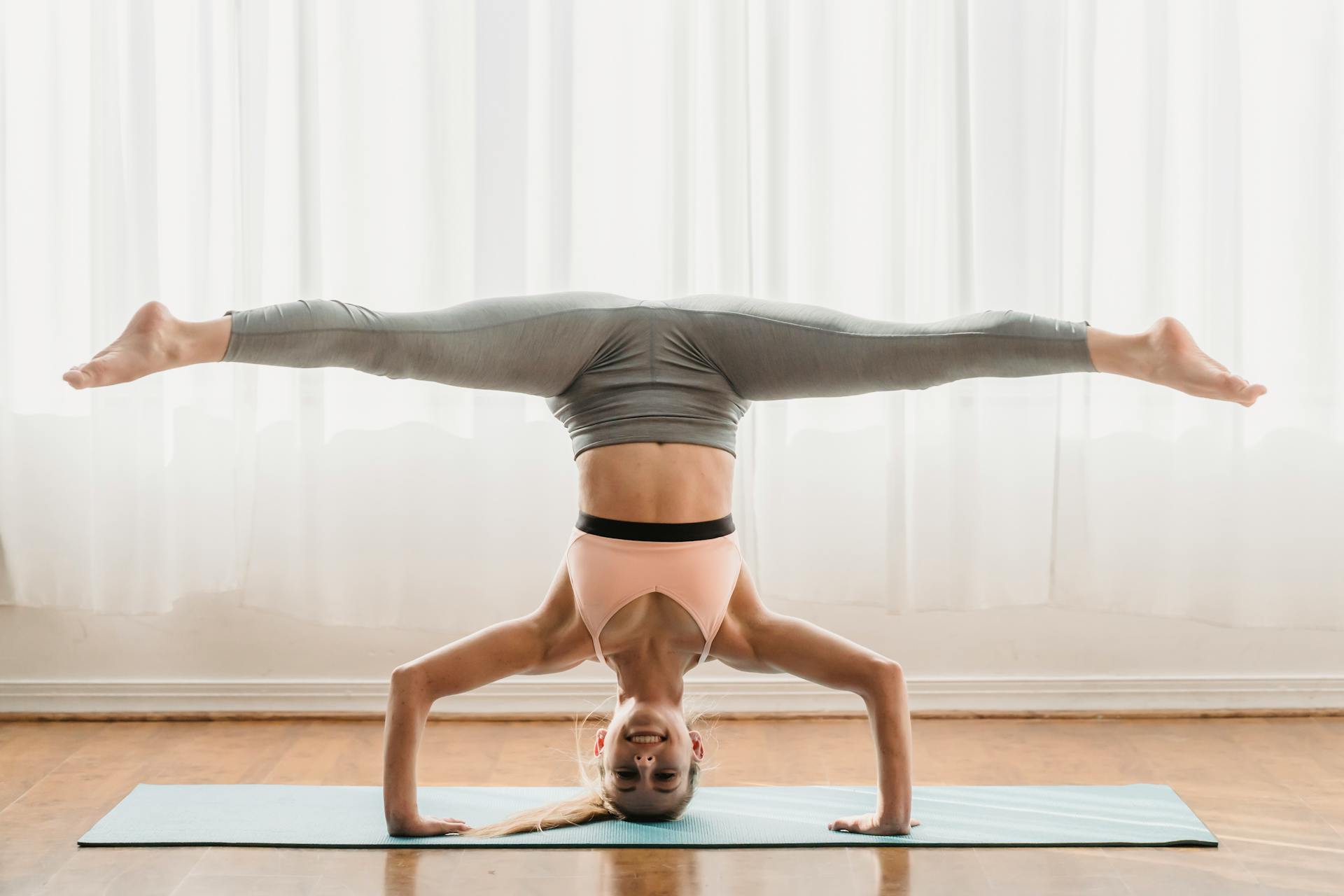 A woman in activewear performs a headstand split yoga pose on a mat indoors, embodying balance and strength.