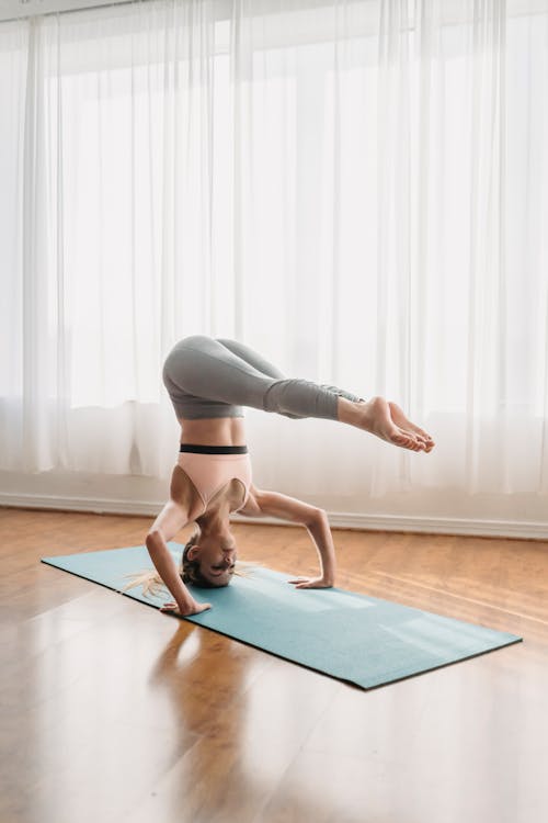 Full length of determined young female in sportswear performing Supported Headstand with Legs Halfway Down on mat during Ashtanga Vinyasa yoga training in light room