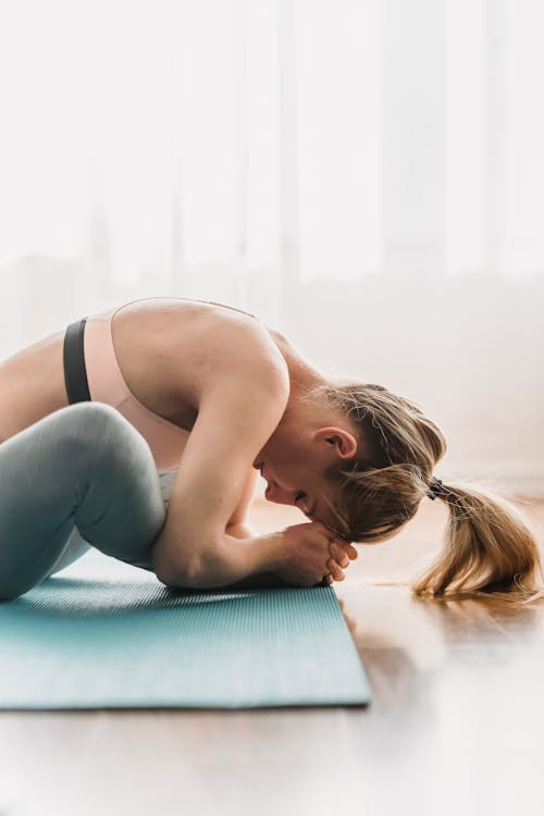 Free Side view of focused young fit woman with long blond hair in sportswear stretching thighs while doing Baddha Konasana B yoga pose with closed eyes in sunny studio Stock Photo