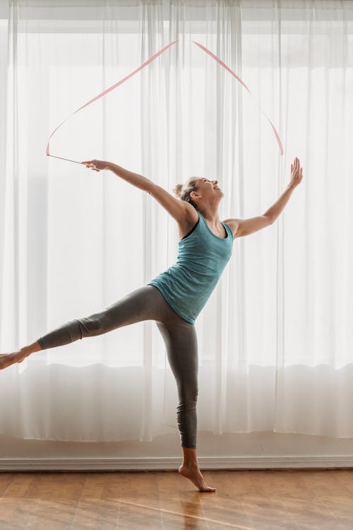 Cheerful young fit female gymnast in sportswear performing dance with ribbon during training in light studio