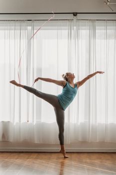 Full length of flexible young feminine lady in activewear performing dance with gymnastic ribbon during rehearsal in studio in sunlight