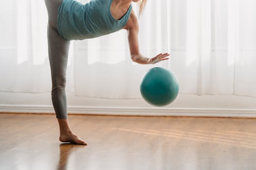 Sportive anonymous barefoot female in activewear standing on one leg while doing exercise with fitness ball during training in room