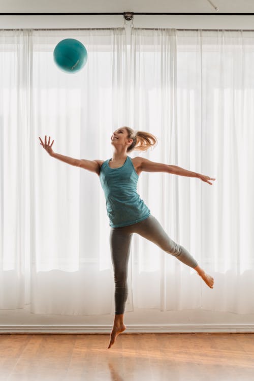 Full body of energetic barefoot female in activewear jumping and throwing fit ball in air while practicing in light room
