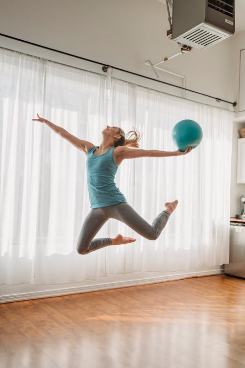 Full body of sportive female in activewear performing gymnastic exercise with fitness ball while training in light room at home