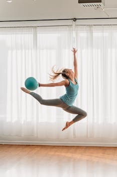 A woman performs an energetic leap with an exercise ball indoors, embodying fitness and flexibility.