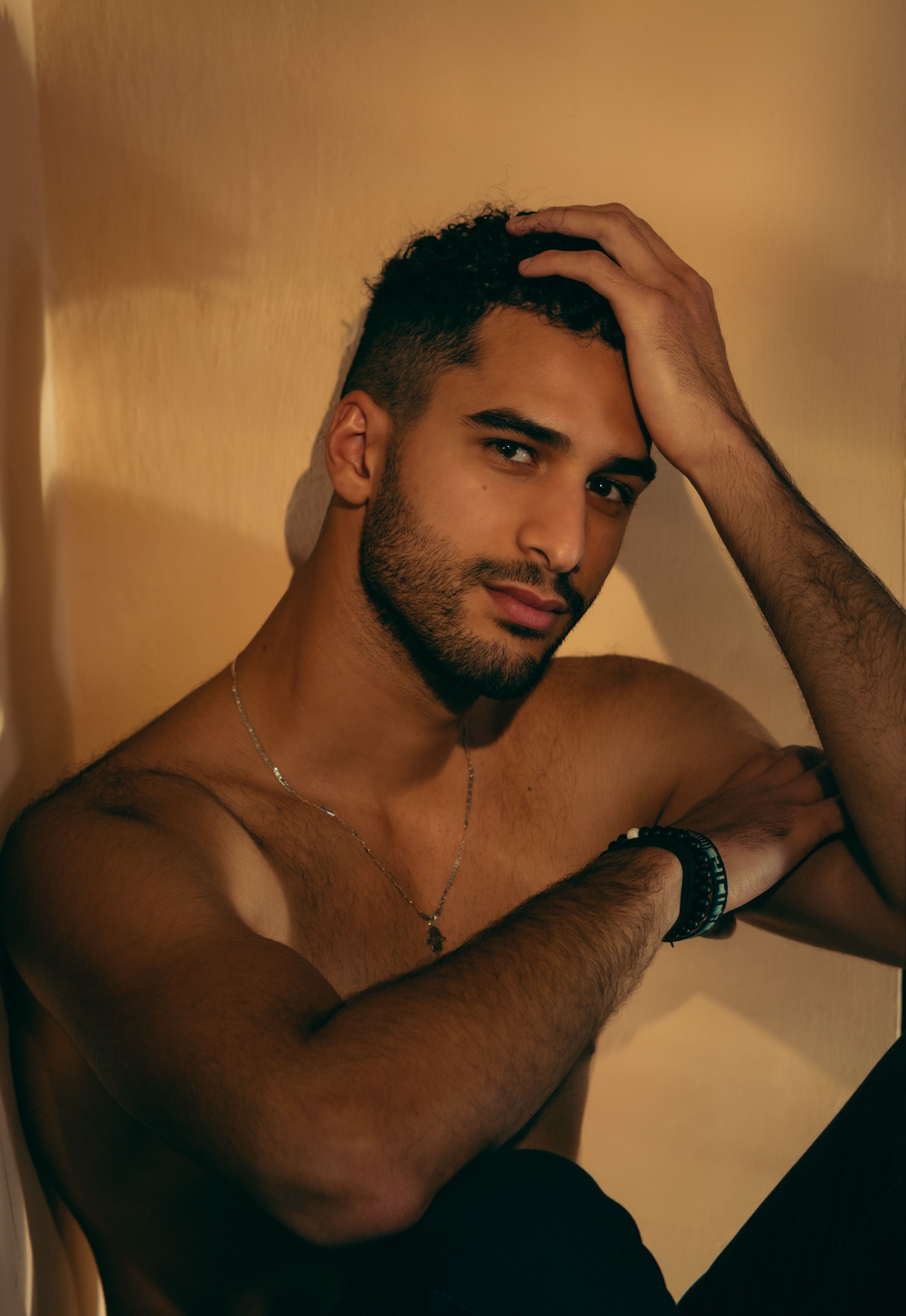 A shirtless man with a contemplative pose under soft indoor lighting, displaying a confident allure.