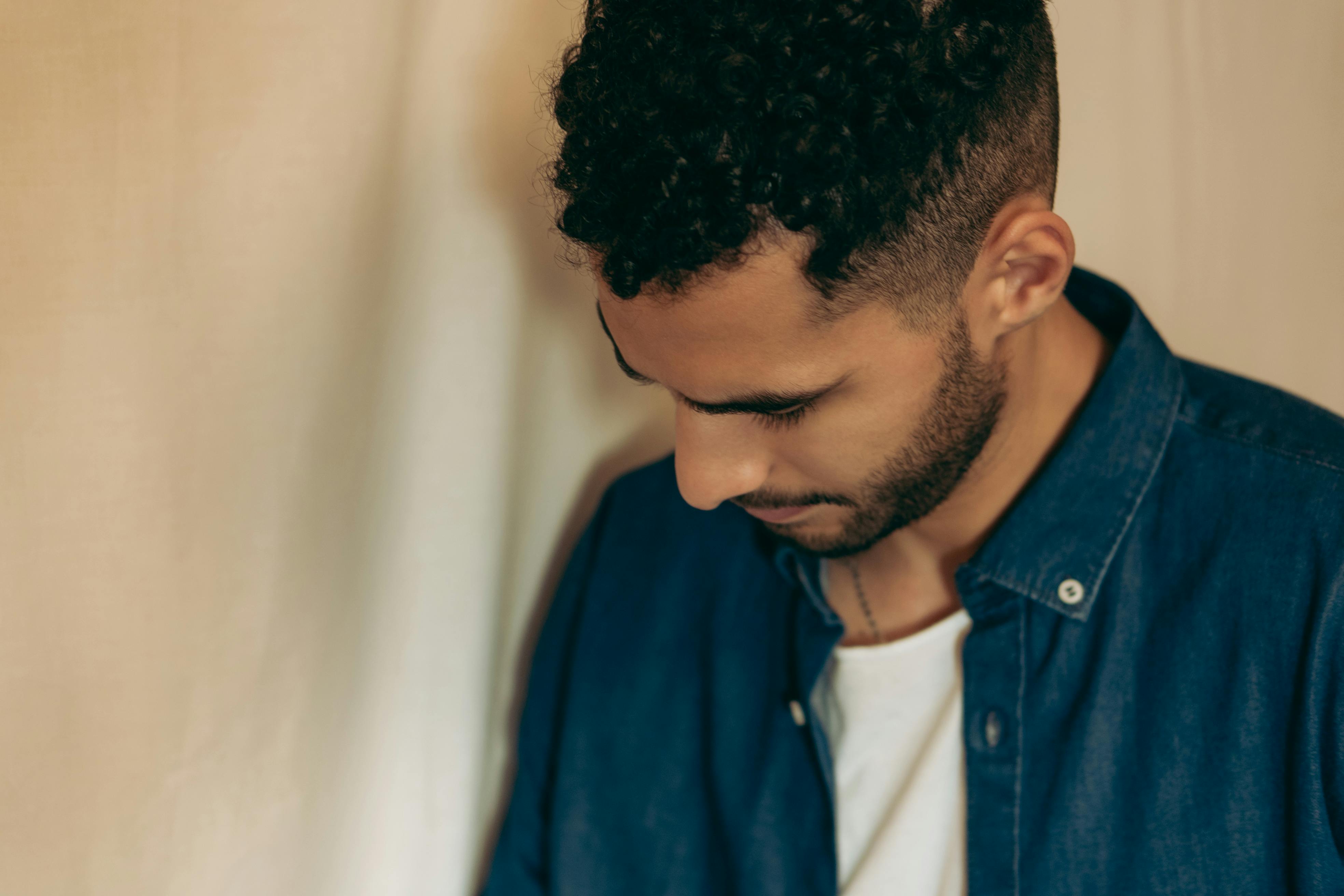 Close-up of a man with curly hair wearing a denim jacket, looking down thoughtfully.