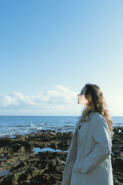 Free Woman in a coat standing by the rocky shoreline, gazing at the ocean under a clear sky. Stock Photo