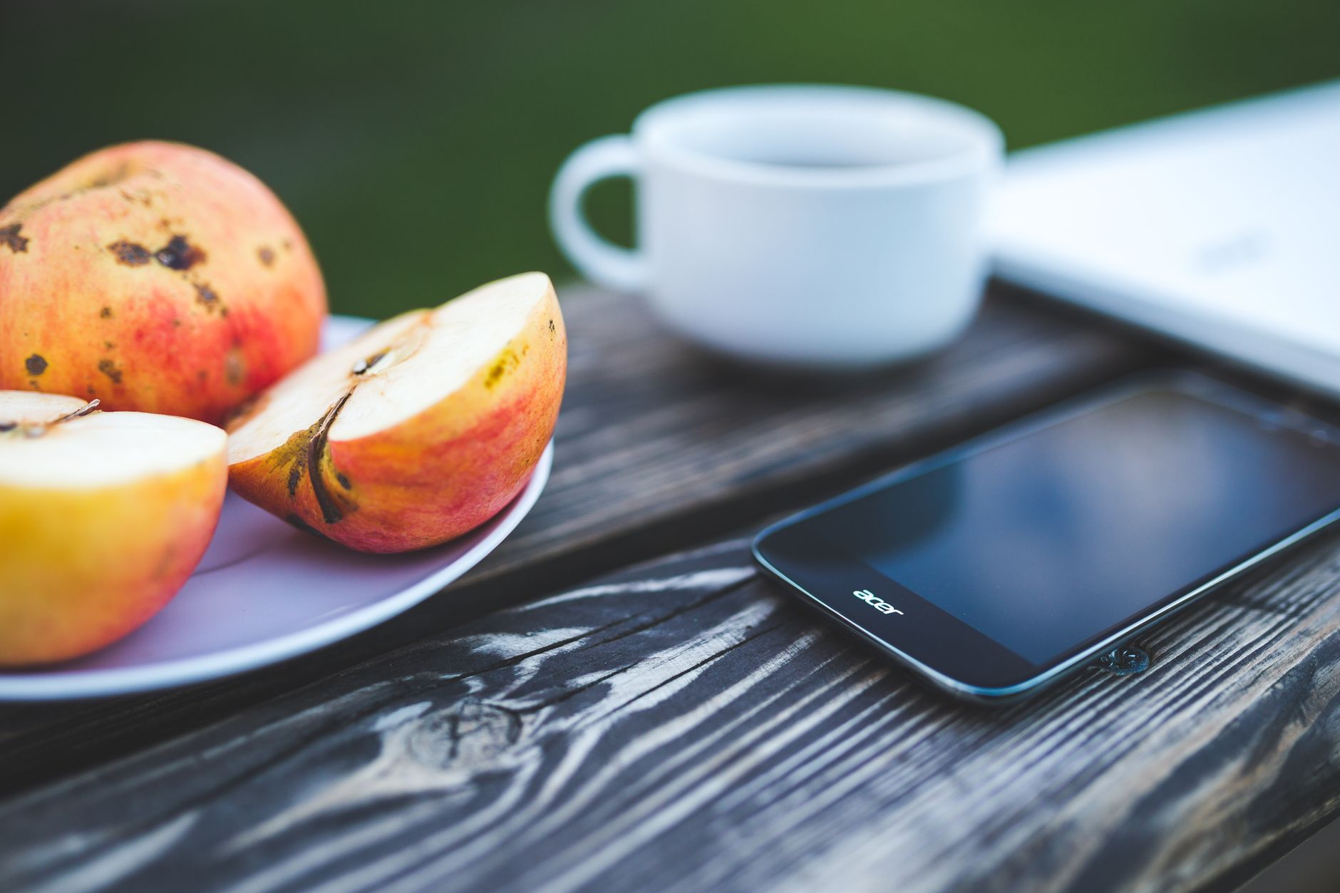 Outdoor scene with sliced apple, smartphone, and coffee on wooden table.