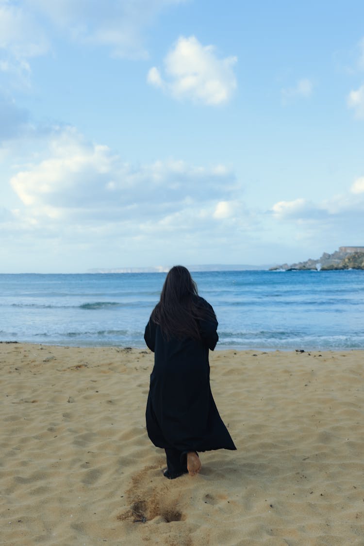 Back View Of A Woman In A Black Coat Walking On Sand