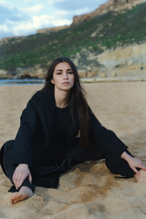 Free Woman in black attire posing on a sandy beach with hills in the background, offering a serene outdoor vibe. Stock Photo