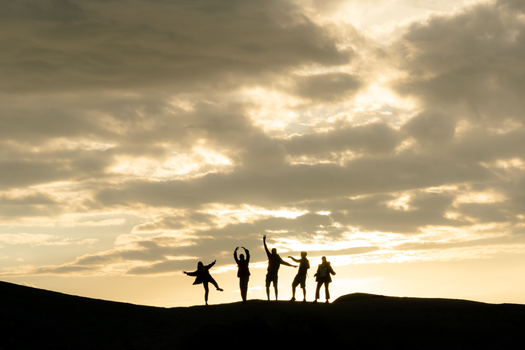 Five silhouetted friends dancing on a hilltop at sunset against dramatic clouds.