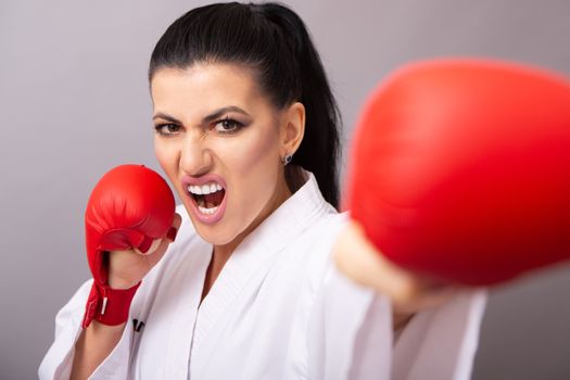Young aggressive female fighter in white sportswear and karate gloves showing hit while looking at camera with open mouth