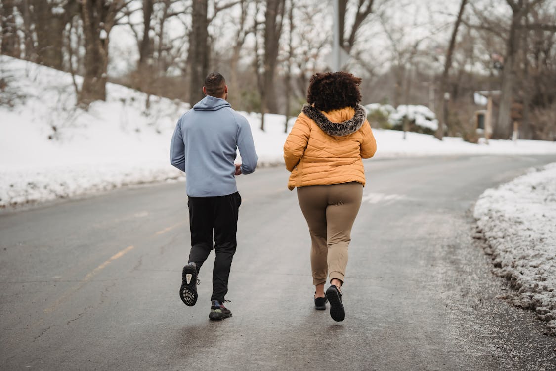 Free Back view full length fit male fitness trainer and plump female in warm jacket jogging together on asphalt roadway in snowy forested suburb on cold winter weather Stock Photo