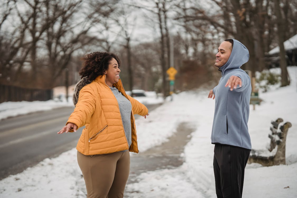 Free Side view happy ethnic fit male trainer and plump black female in warm clothes performing side arms raise exercise while warming up on snowy winter sidewalk Stock Photo