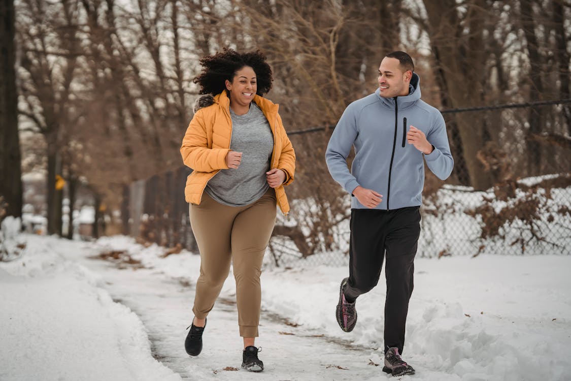 Free Full length smiling ethnic male fitness trainer and plus sized black female in warm jacket jogging together on snowy pathway in frozen winter park Stock Photo