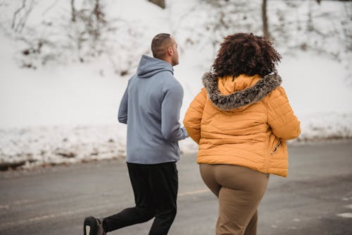 Back view unrecognizable ethnic sporty male trainer and plump female in outerwear jogging together on snowy roadside on cold winter weather