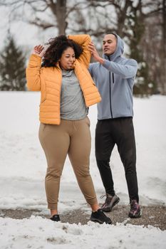 Full length professional ethnic male trainer in activewear helping plus sized black female to stretch arm behind head while working out in snowy winter park