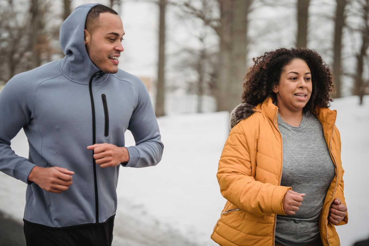Plus-size woman in a mustard puffer running beside a trainer in a snowy park