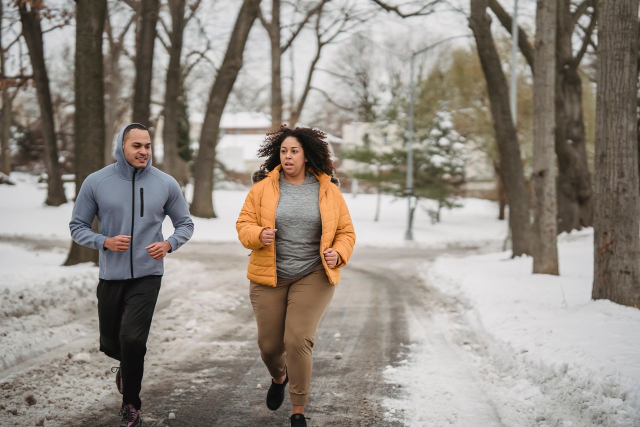 Plus-size woman in a mustard puffer jacket running with a trainer in a snowy park