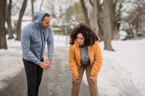 Tired plus size African American female with black personal instructor standing on pathway during training in winter park on blurred background