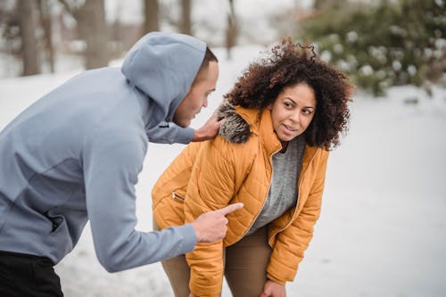 Black personal trainer pointing away and touching shoulder of plump African American female during training in snowy park on blurred background