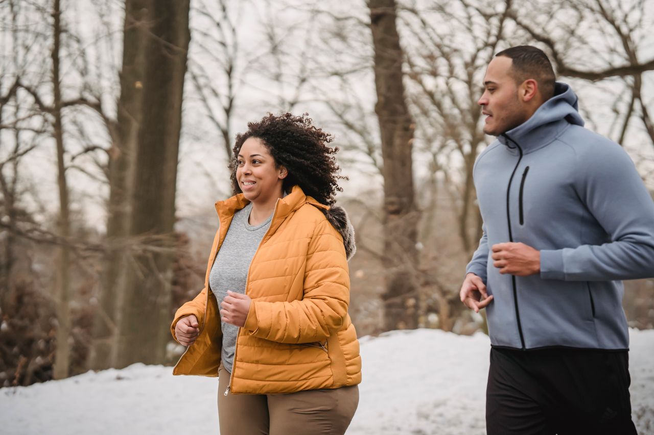Plus-size woman in a mustard puffer running through a snowy park with a trainer