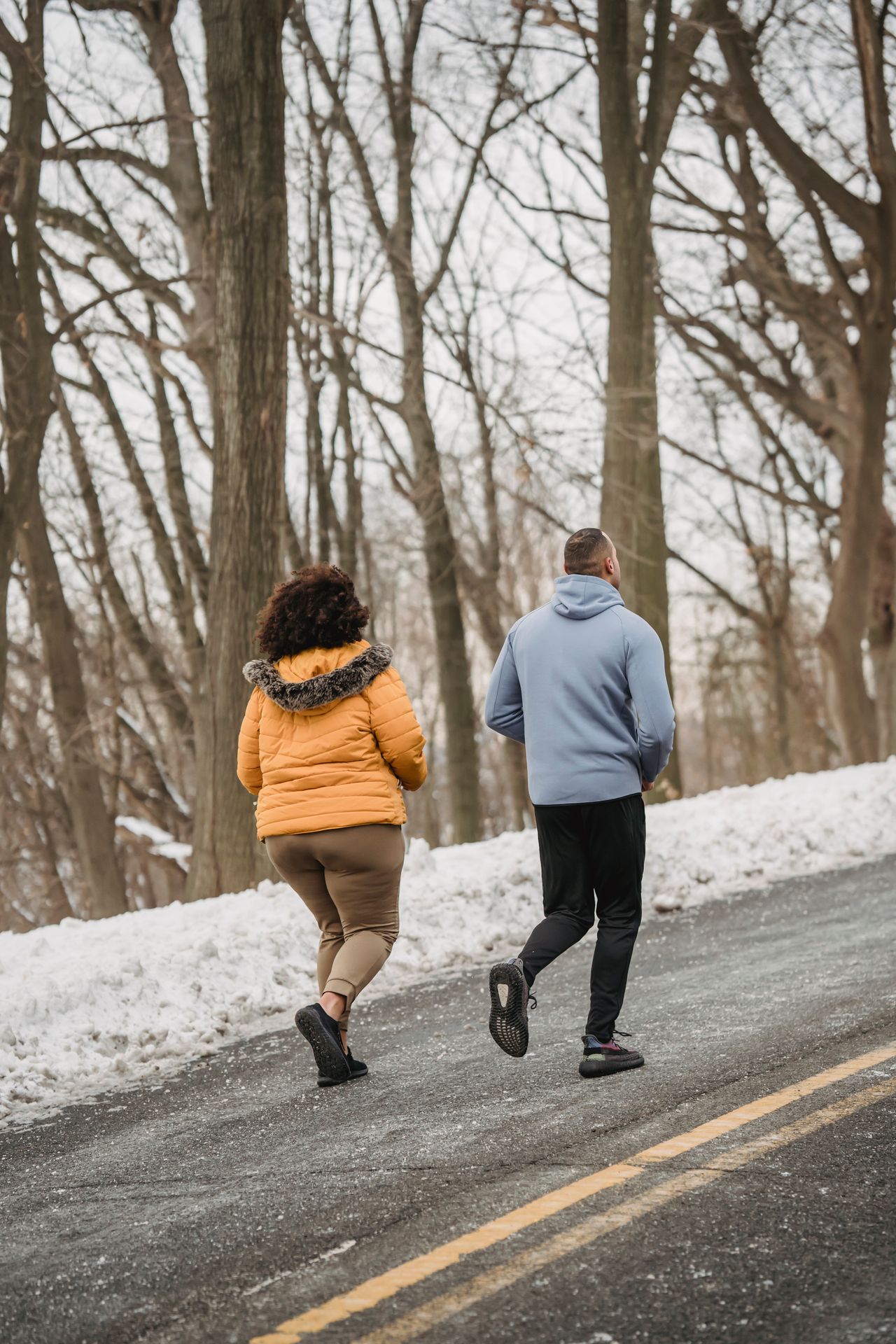 Back view of a plus-size woman in a mustard puffer running with a trainer on a snowy road