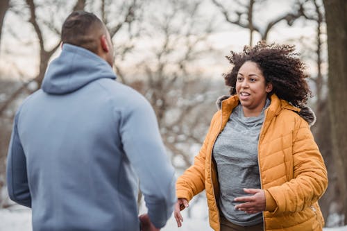Free Positive plump African American in warm jacket looking at anonymous personal coach while standing in snowy nature with trees during training Stock Photo