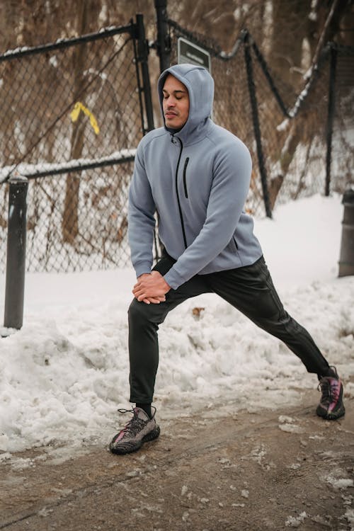 Full body of determined African American male wearing sportswear doing stretching exercise while standing on snowy pathway near fence during training