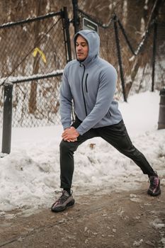Full body of determined African American male wearing sportswear doing stretching exercise while standing on snowy pathway near fence during training