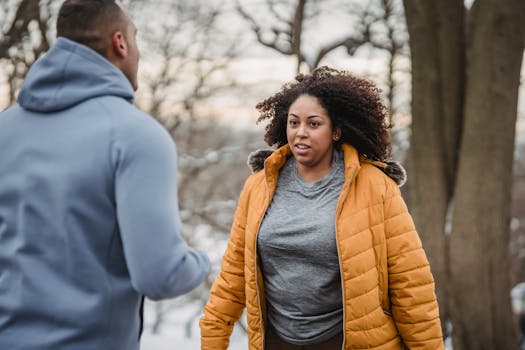 Plump African American female in warm outerwear with unrecognizable personal trainer standing in nature with trees during workout in winter time