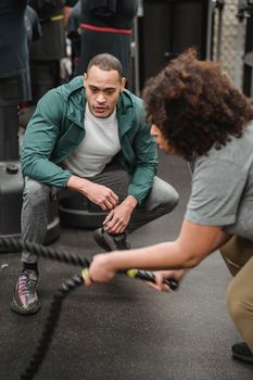 Dedicated ethnic female in sportswear exercising with battling ropes near fit professional male trainer in modern gym