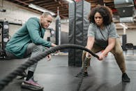 Full body determined African American female in activewear exercising with battling ropes near serious male fitness instructor in contemporary sports club