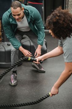 Determined black woman exercising with battle ropes near hunkering fit male instructor during functional workout in gym