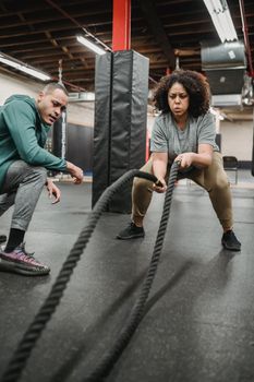 Strong African American female in activewear training with battle ropes near fit male coach in fitness center