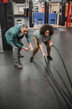 Full body focused African American female in activewear performing battle ropes exercise near professional male instructor in modern fitness center
