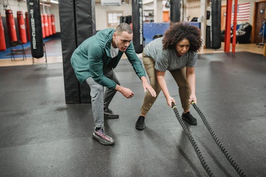 Full body focused African American female in activewear exercising with battle ropes near professional male instructor in contemporary fitness center