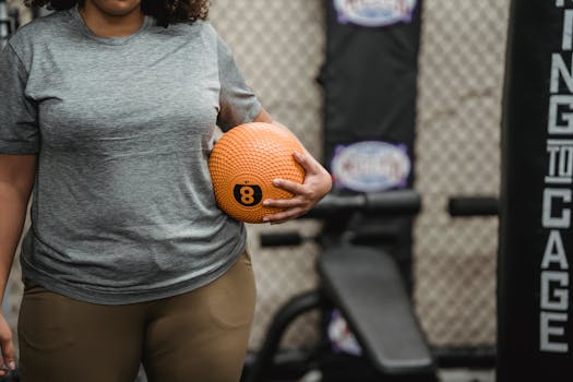 Focused image of a plus-size woman holding an orange medicine ball in a gym setting.