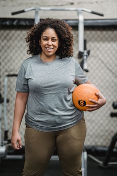Positive young plus size African American female with curly hair in activewear smiling and looking at camera while standing in modern gym with medicine ball in hand