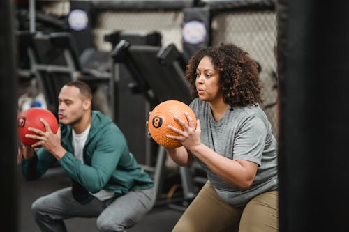 Free Focused young plus size black female in sportswear doing squats with medicine ball while training in gym with muscular ethnic male trainer Stock Photo
