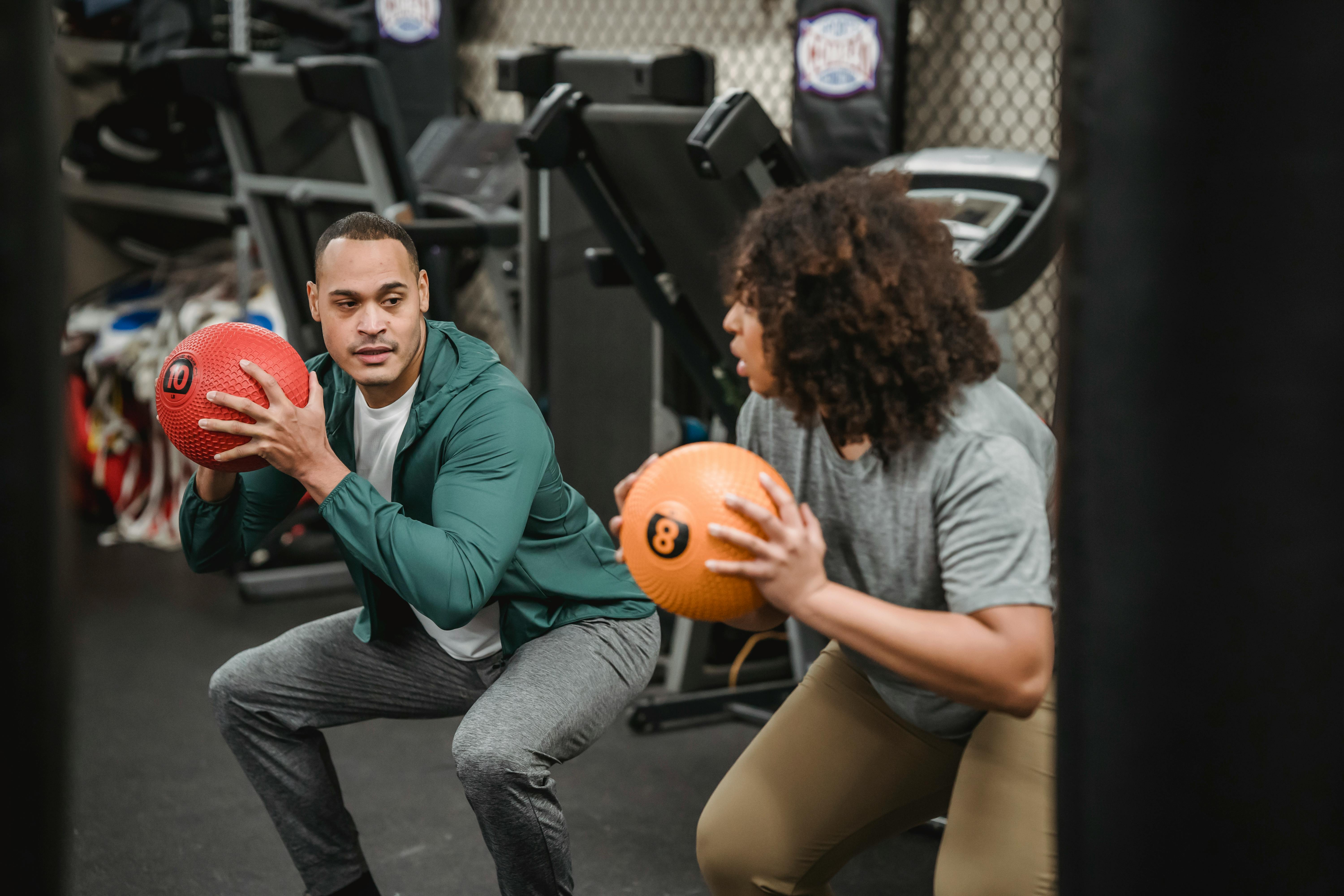 Concentrated young multiethnic male instructor and female client doing butt exercise with medicine balls and looking at each other during training in gym