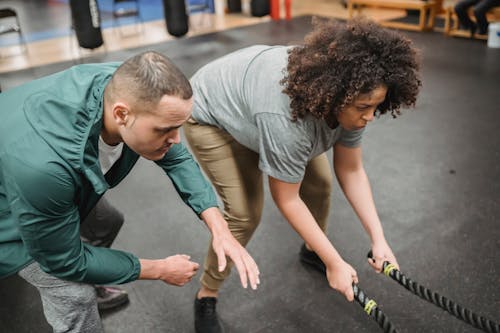Free Focused young male instructor in sportswear helping overweight African American female exercising with battle ropes in modern gym Stock Photo
