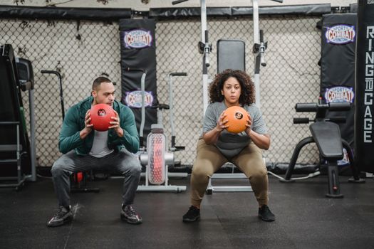 Full body of strong young ethnic male trainer and plus size black female client in sportswear doing static squat exercise with medicine balls during functional workout in gym