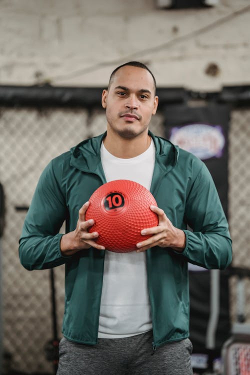 Confident young ethnic male athlete in sportswear holding medicine ball in hands and looking at camera while training in gym