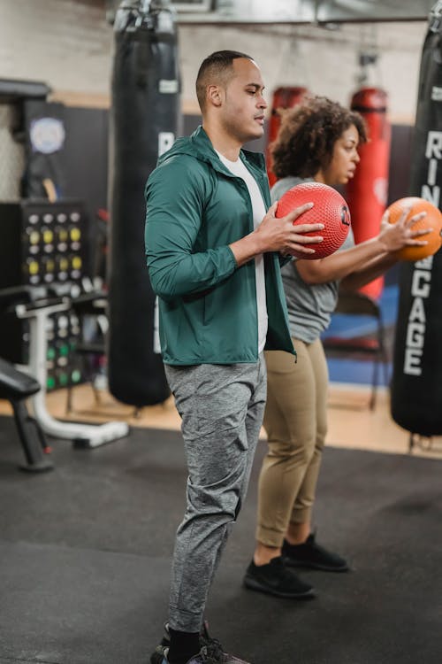 Side view of calm young diverse male and female in sportswear exercising with weight balls while standing together in modern gym