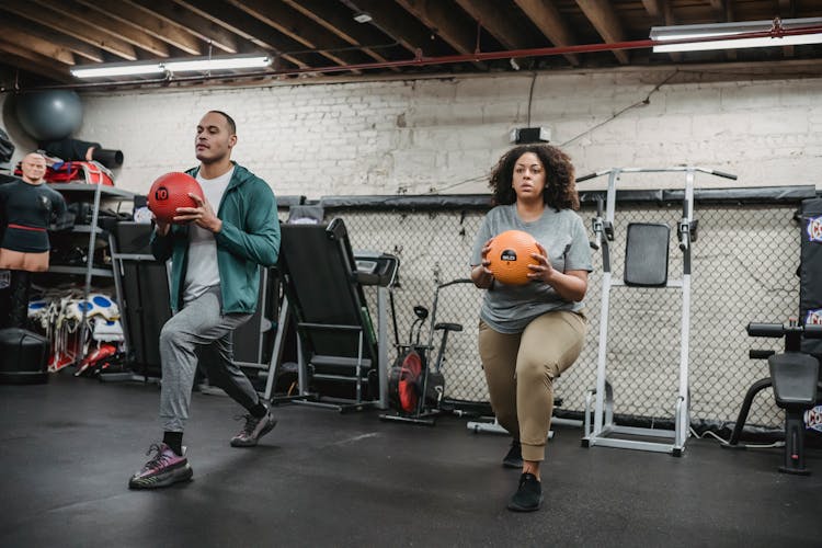 Young Multiracial Man And Woman Doing Lunges Exercise With Weight Balls In Gym