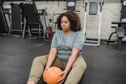 A plus size woman takes a break at the gym, holding an exercise ball, symbolizing determination and wellness.