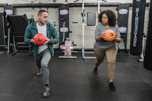 Full body of determined plus size black woman in activewear doing lunges with weigh ball during functional workout with ethnic male personal trainer in sport club