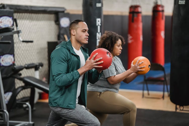 Strong Multiracial Trainer And Sportswoman Doing Lunges With Balls In Gym